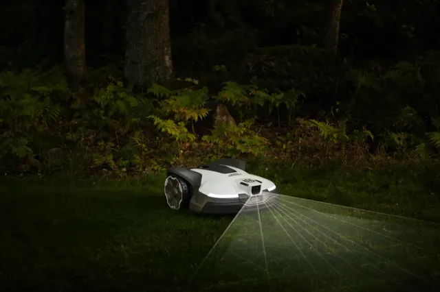 Robotic lawn mower operating in a dimly lit garden with laser guidance lines visible.